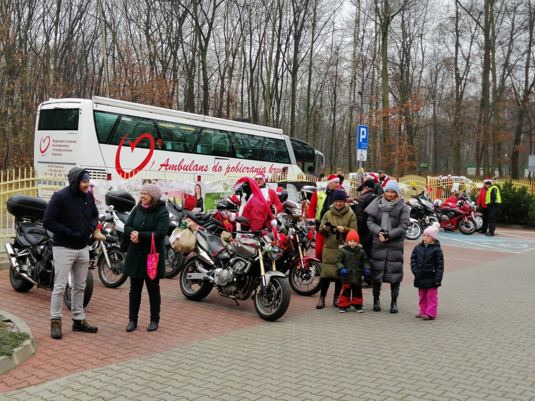 Dorośli i dzieci stojący i uśmiechający się. Stoją na parkingu, obok nich zaparkowane motocykle. Za nimi stoi autobus amnulans do pobierania krwi