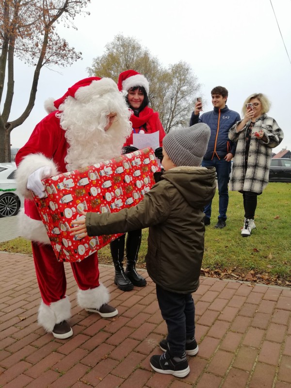Mikołaj wręcza duży prezent dziecku. z Tyłu dwie osoby robią zdjęcia i nagrywają filmy telefonami komórkowymi