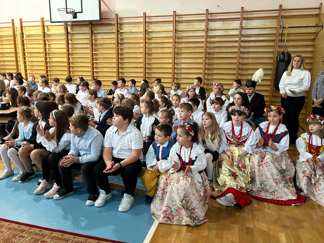 Grupa dzieci siedzi na sali gimnastycznej i obserwuje wydarzenie