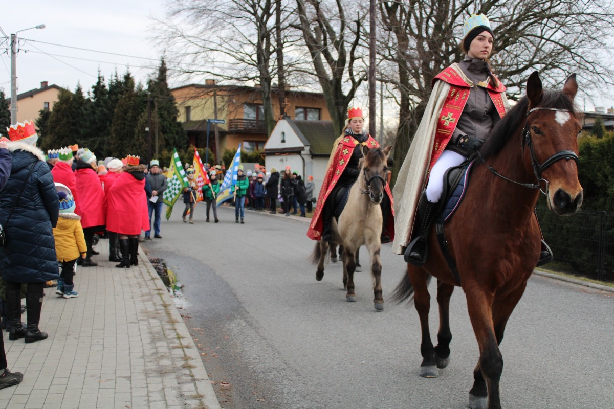 Dwie kobiety jadą konno. za nimi idą Ludzie w koronach na głowie idą ulicą podczas orszaku trzech Króli