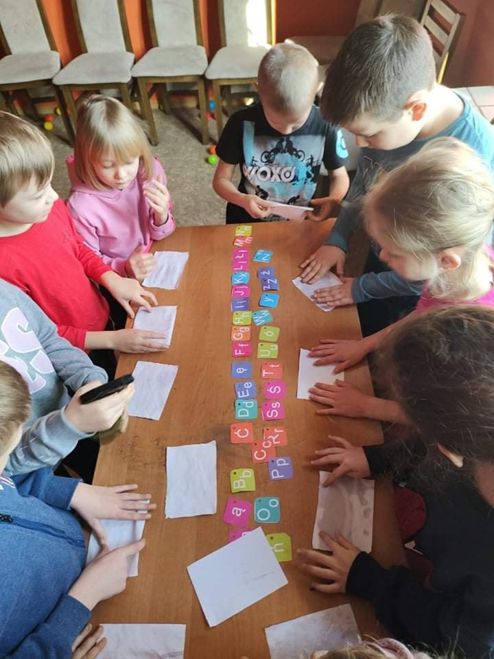 Dzieci podczas zabawy detektywistycznej stoją przy stole, na stole kolorowe kartki z literami