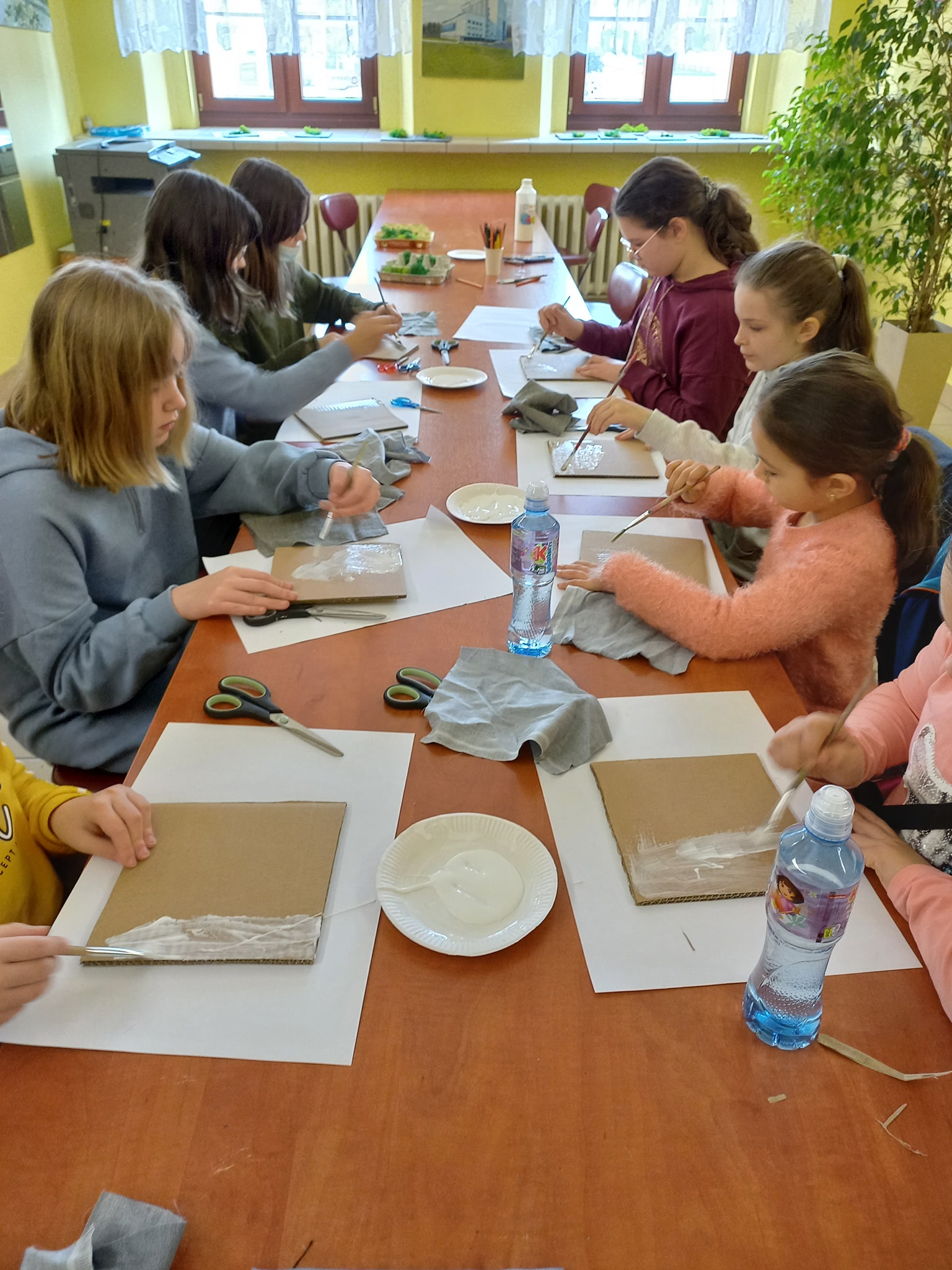 Dzieci przygotowują prace plastyczne siedząc przy stole. Na stole klej, nożyczki i kartoniki na których przygotowywane są prace