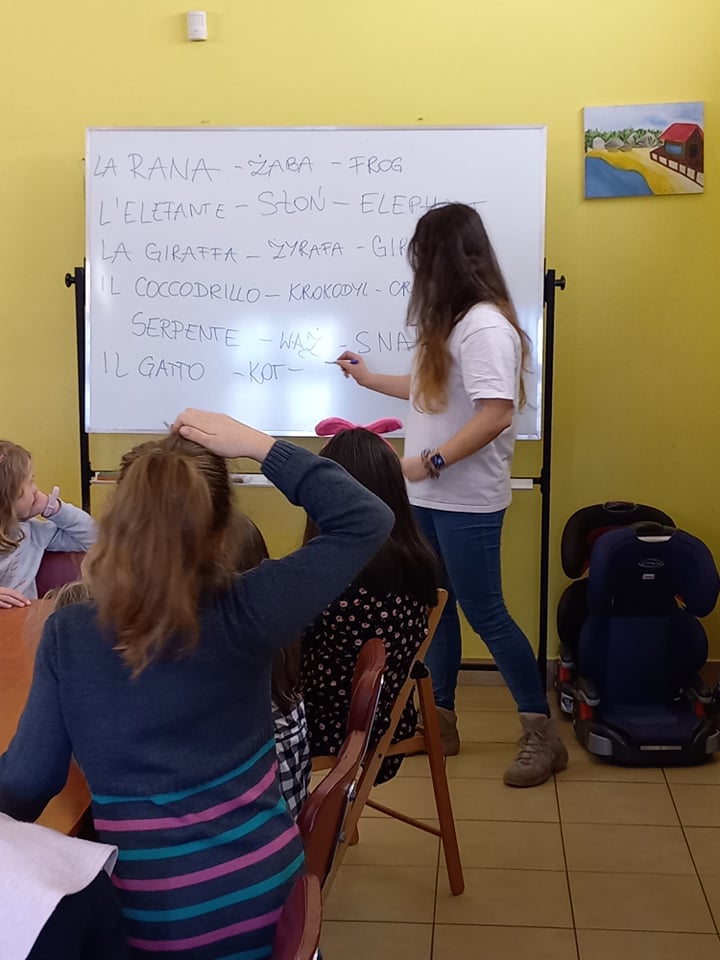 zajęcia językowe, nauczycielka stojąca przy tablicy, dzieci siedzące przy stołach patrzą na tablicę