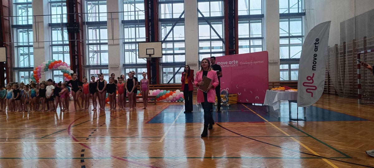Grupa młodych akrobatów w strojach gimnastycznych stoi na parkiecie hali sportowej, czekając na rozpoczęcie wydarzenia. W tle znajduje się dekoracja z balonów w pastelowych kolorach oraz różowe stoisko z logotypem „Move Arte”. Na pierwszym planie kobieta