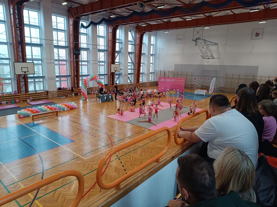 Widok z trybun na halę sportową, gdzie trwa pokaz akrobatyczny. Młodzi uczestnicy w różowych strojach wykonują układy na specjalnych matach, a publiczność z zainteresowaniem obserwuje występy. Dekoracje z balonów i stoisko organizatora tworzą kolorowe tło