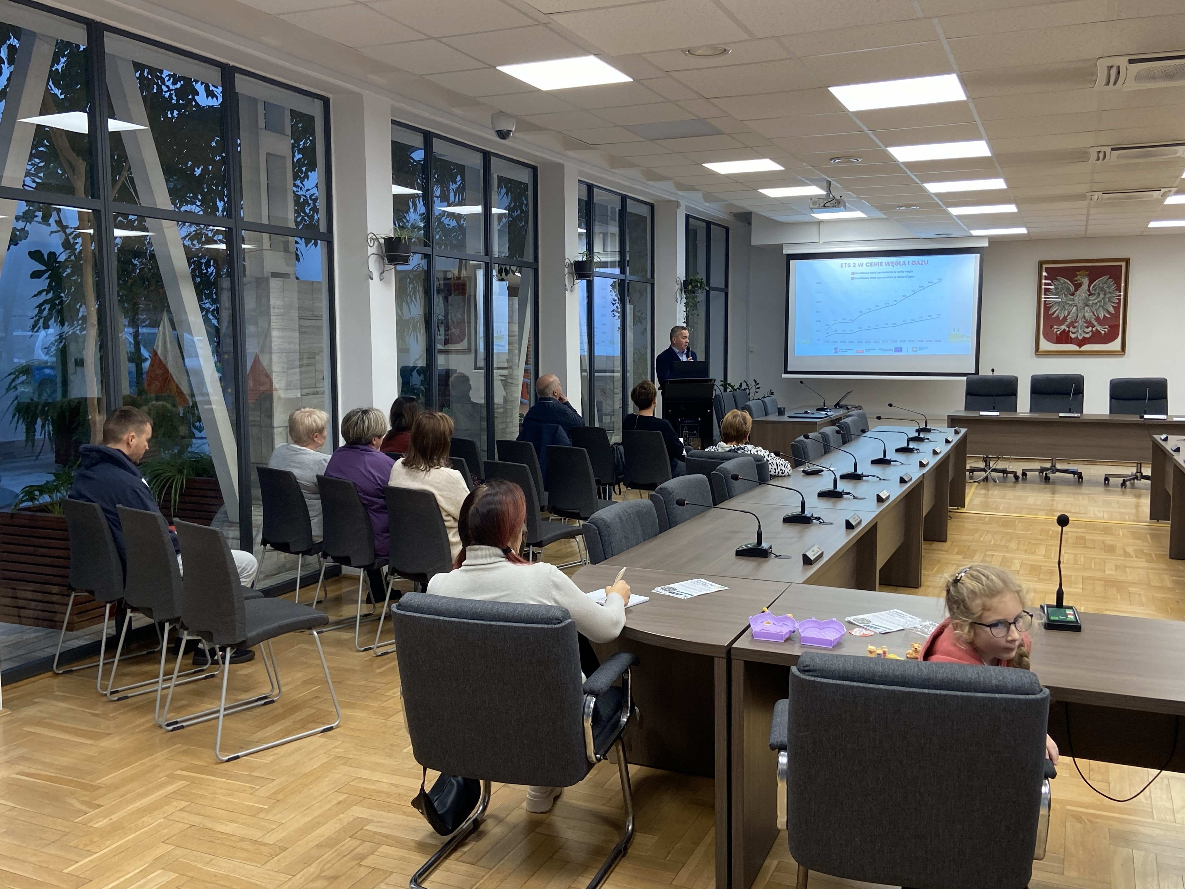 Widok ogólny sali konferencyjnej. Grupa kilkunastu osób siedzi, słuchając prelegenta stojącego z tyłu obok rzutnika. Na ekranie widoczny wykres. Na ścianie herb Polski.